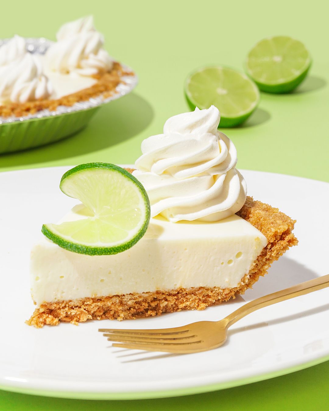 Key lime pie slice with whipped cream and lime garnish on white plate against green background.