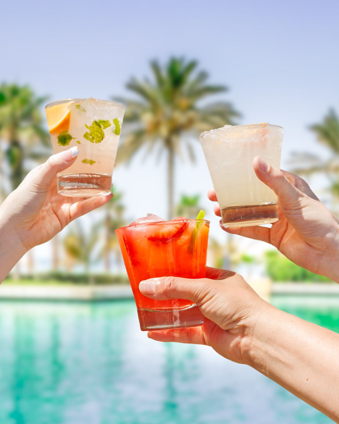 Hands holding colorful tropical cocktails by a resort pool with palm trees in background.