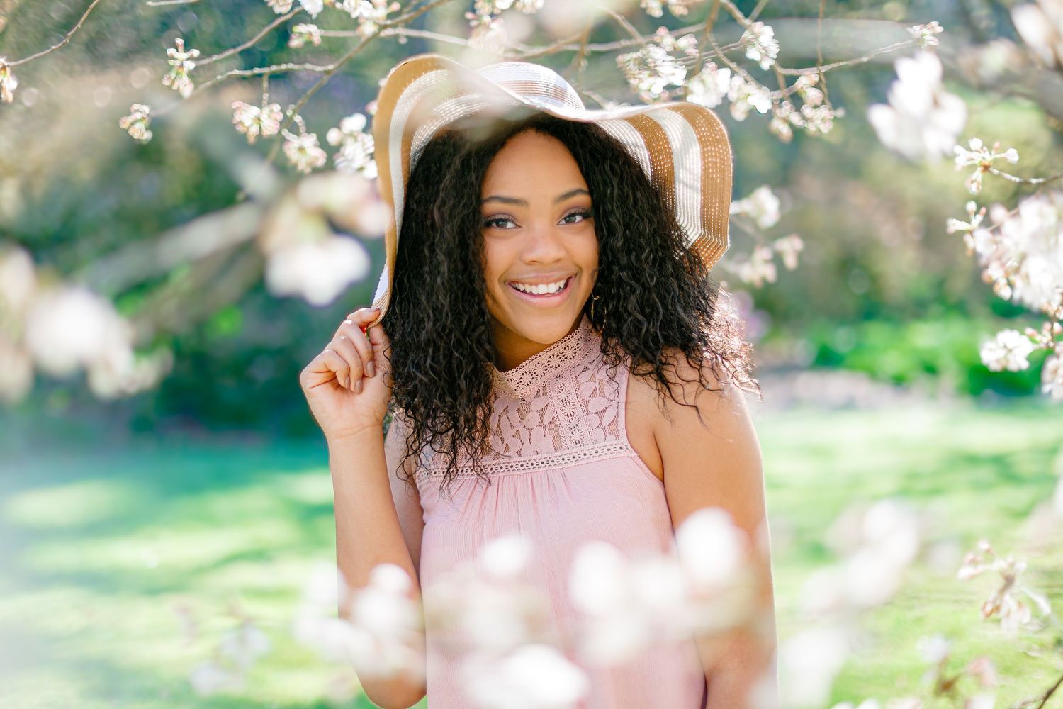 Woman in pink lace dress and striped sun hat poses among spring cherry blossoms.