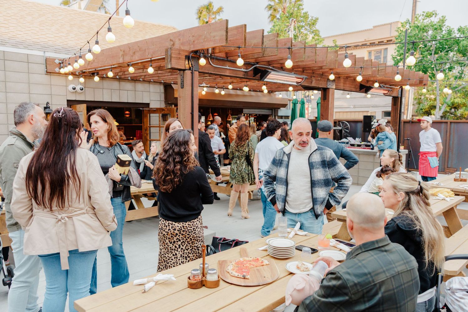 Lively outdoor dining scene with guests mingling under wooden pergola and string lights.