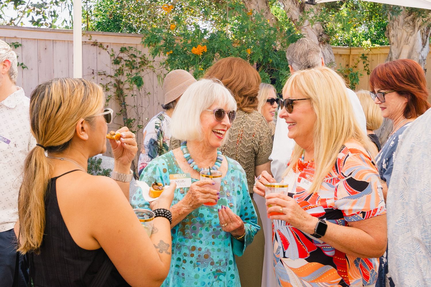 A lively outdoor garden party with guests mingling and socializing under trees and umbrellas.