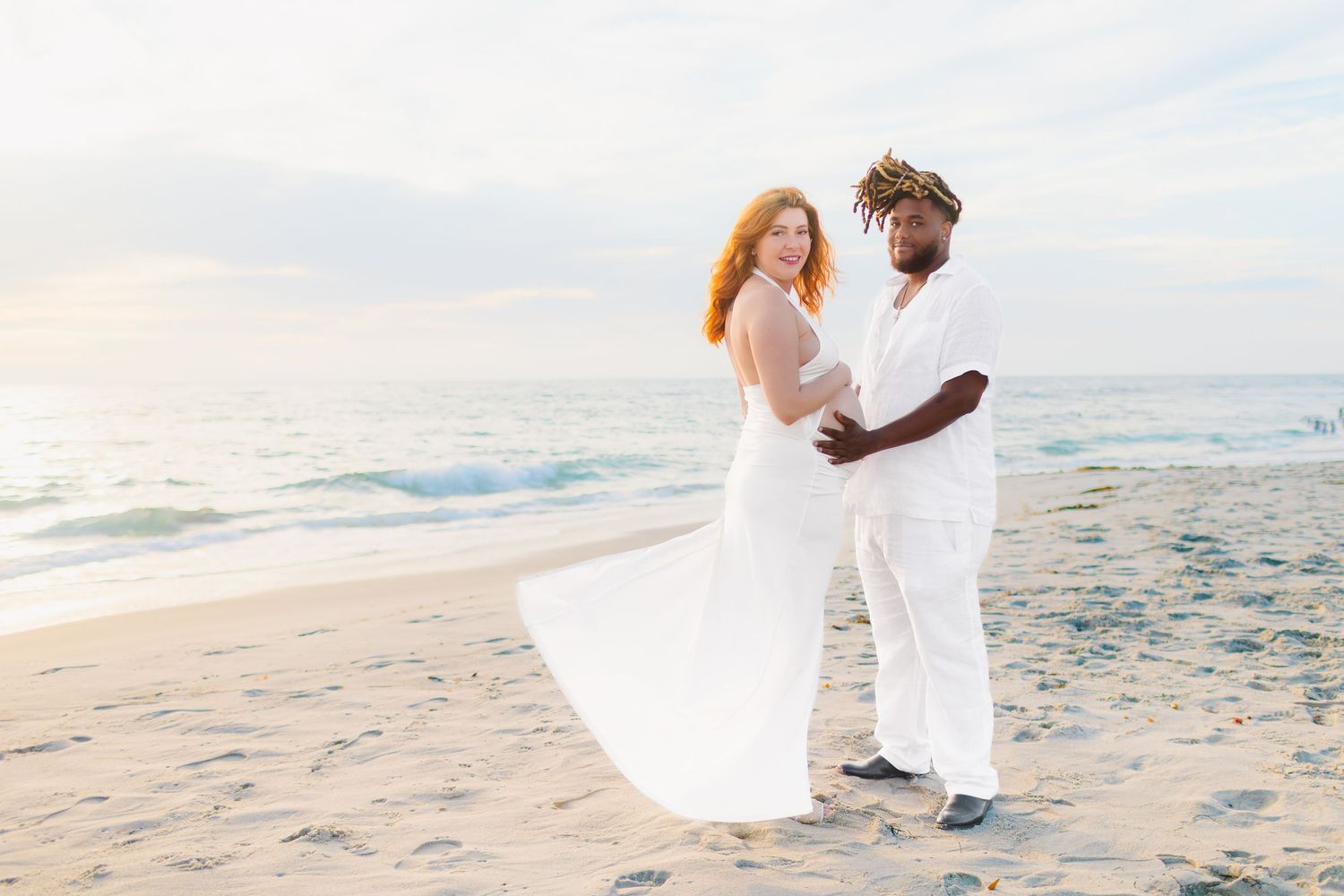 A couple in white attire shares romantic moments on a sandy beach during sunset.
