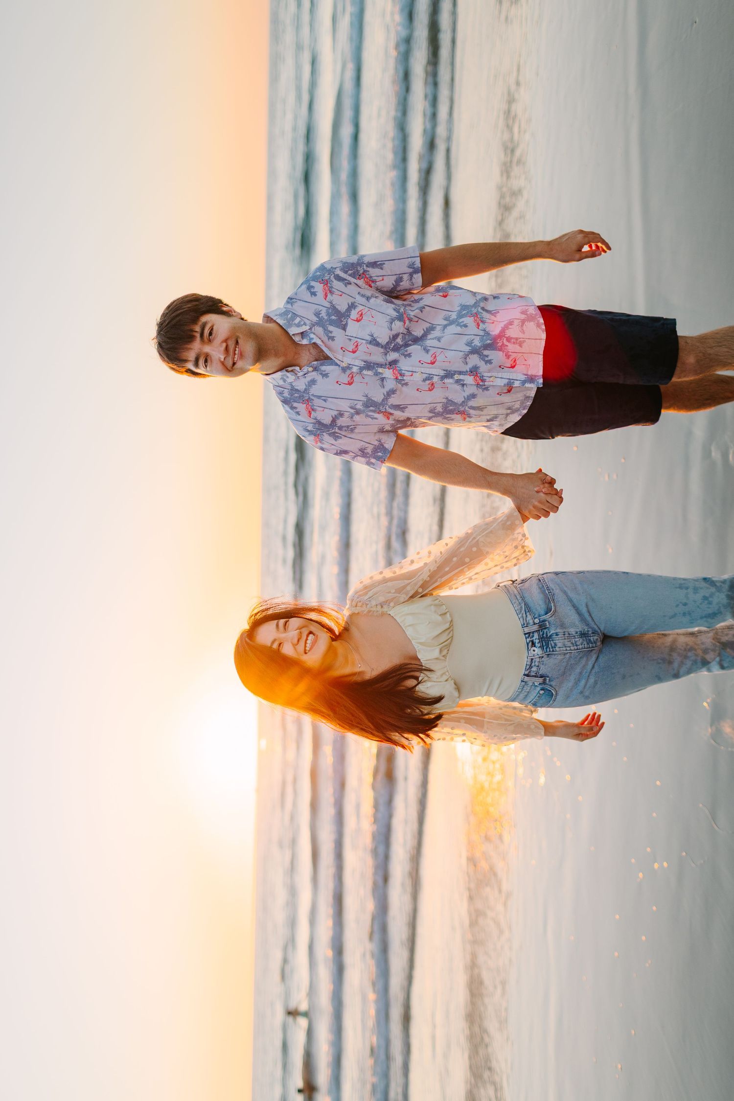 Couple poses together on beach at sunset with soft orange glow and ocean waves behind them.