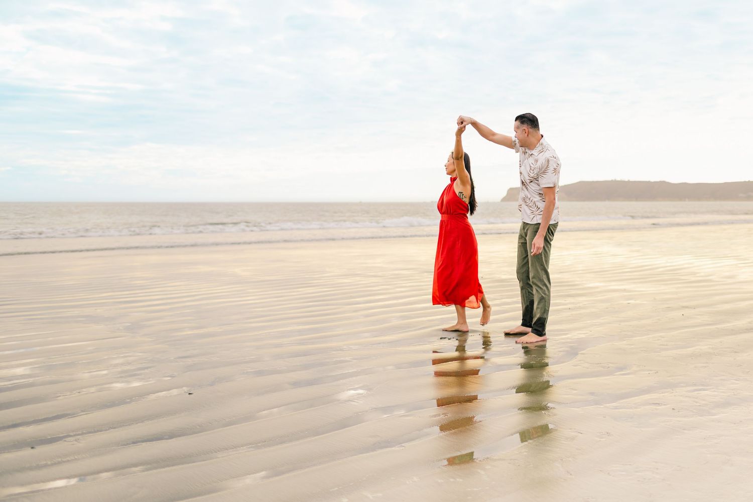 A couple stands together on a sandy beach during a romantic photoshoot with the person in a flowing red dress.