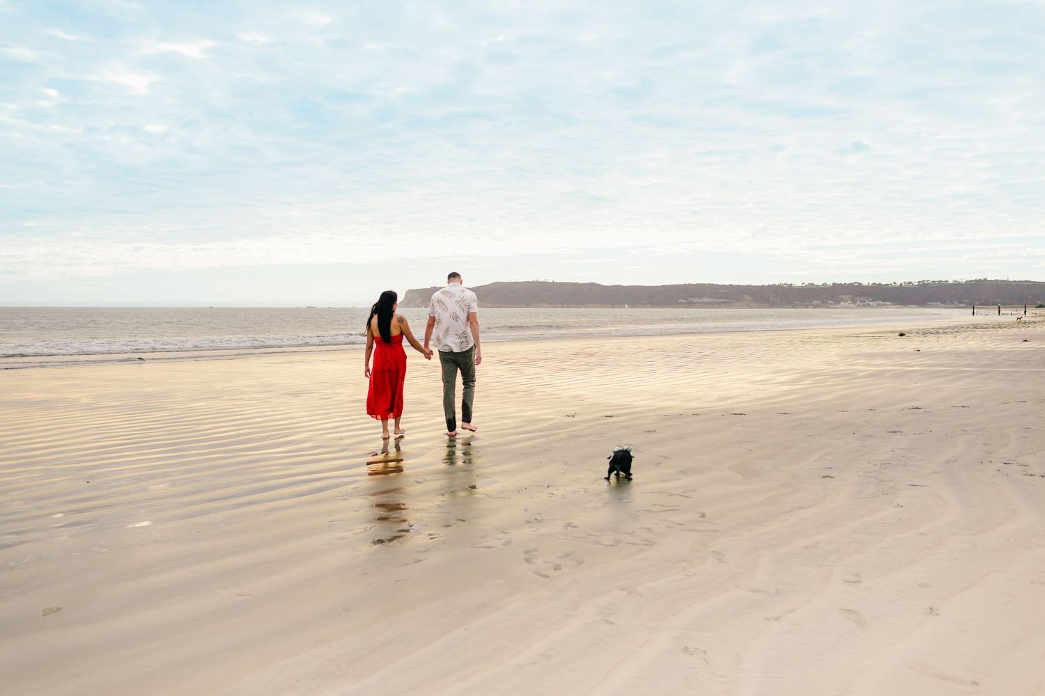 A couple and their dog walking along a vast empty beach during sunset with soft golden light.