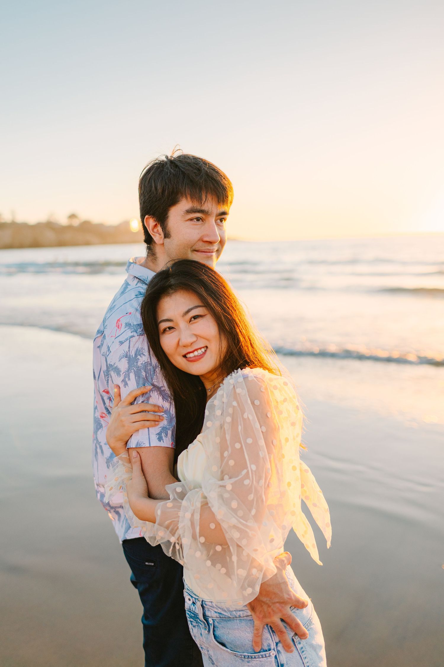 Romantic couple embraces on beach at sunset while wearing white lace and blue floral outfits.