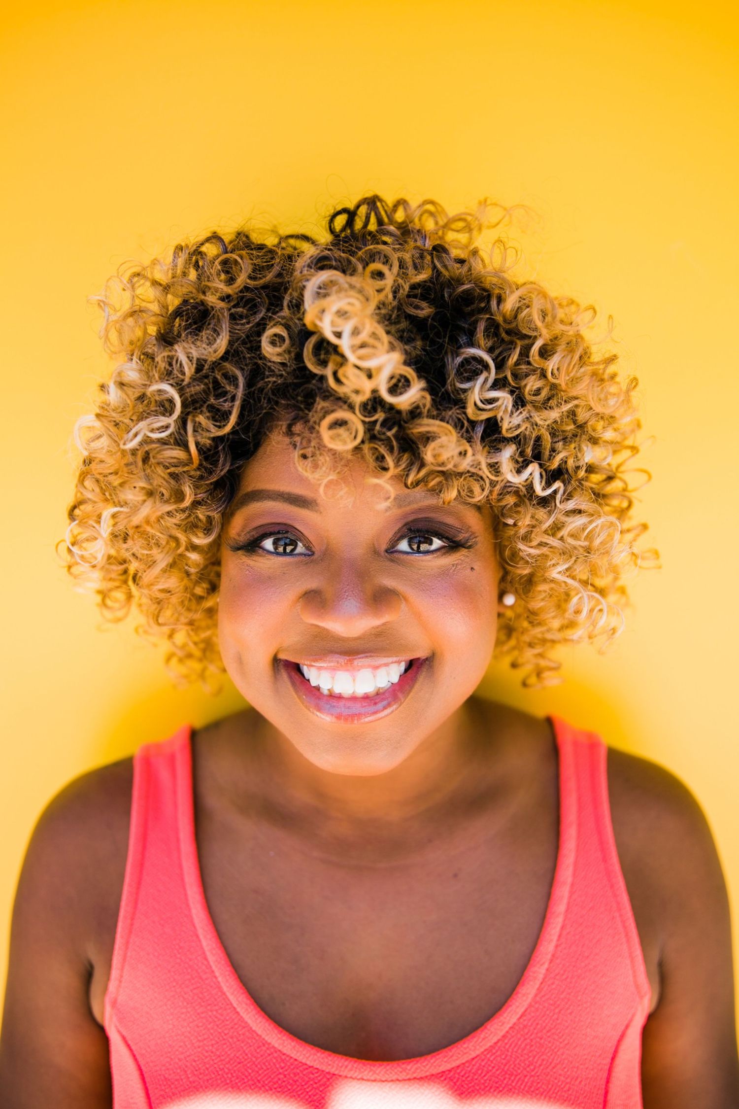 Woman with curly blonde highlights smiling against bright yellow background wearing coral tank top.