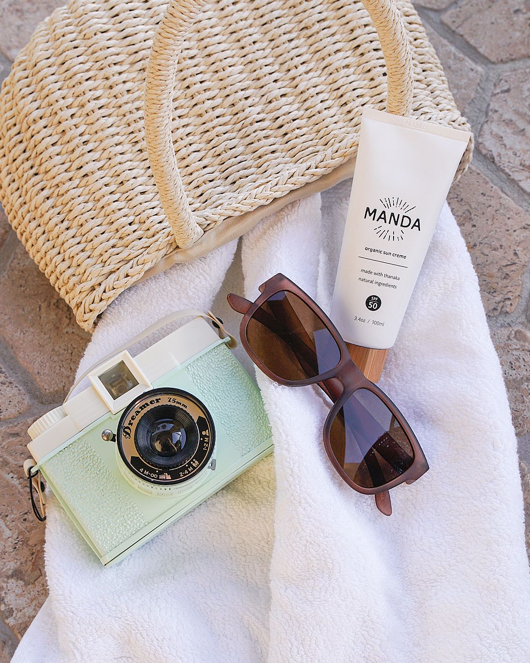 Vintage camera, sunglasses and Manda products arranged on white towel and woven texture.