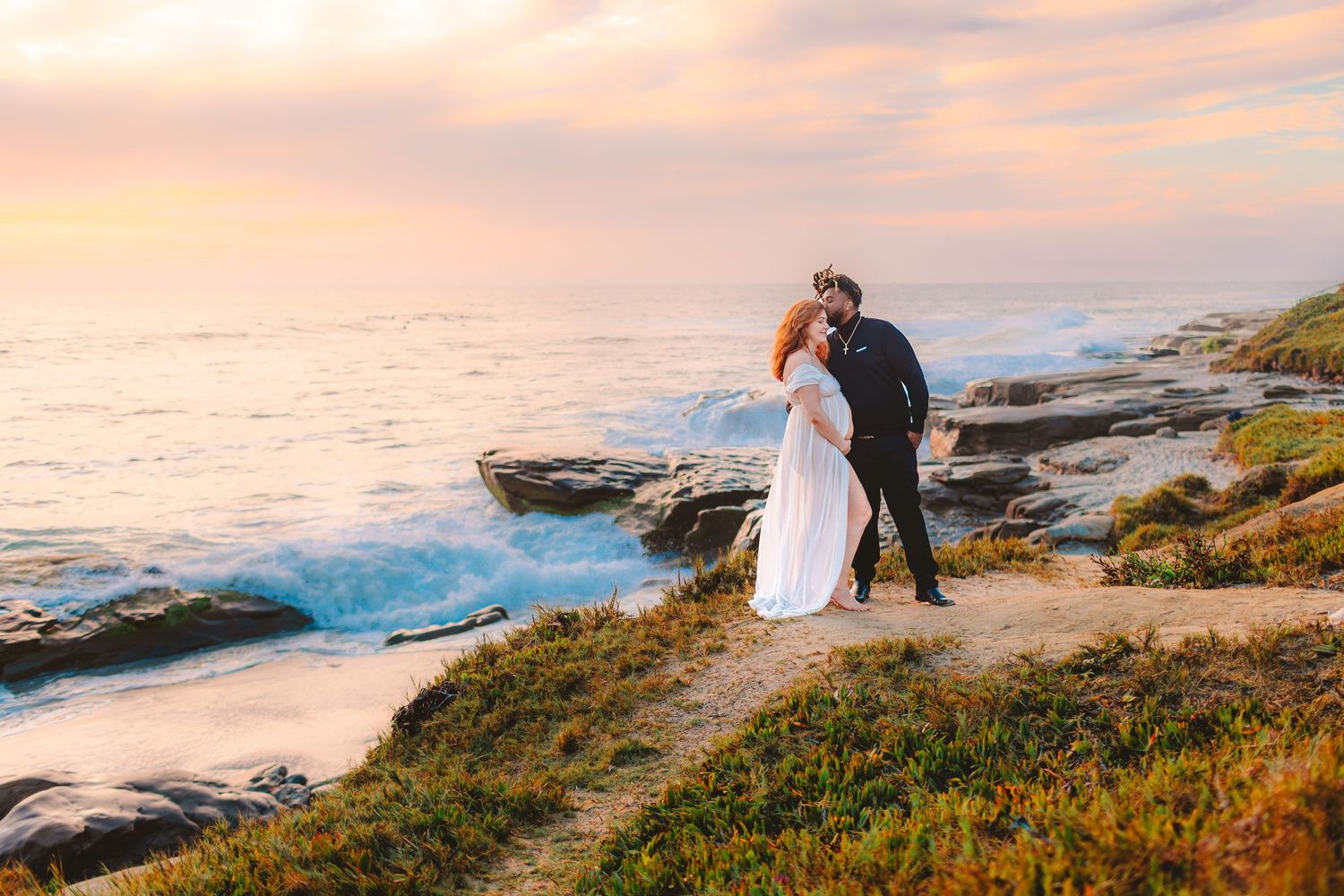 A couple in wedding attire stands on rocky coastal cliffs at sunset overlooking crashing waves and ocean views.