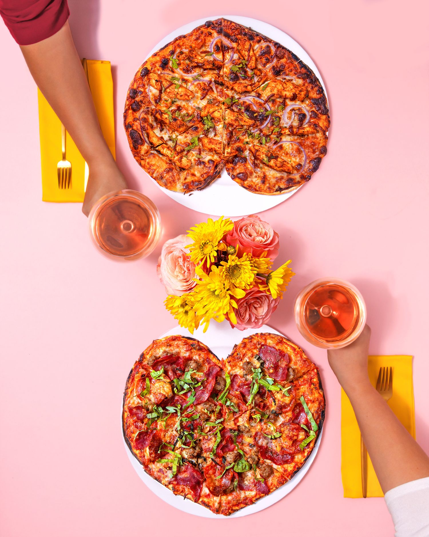 Overhead view of spicy pepperoni pizzas and wine glasses on a pink background with yellow napkins.
