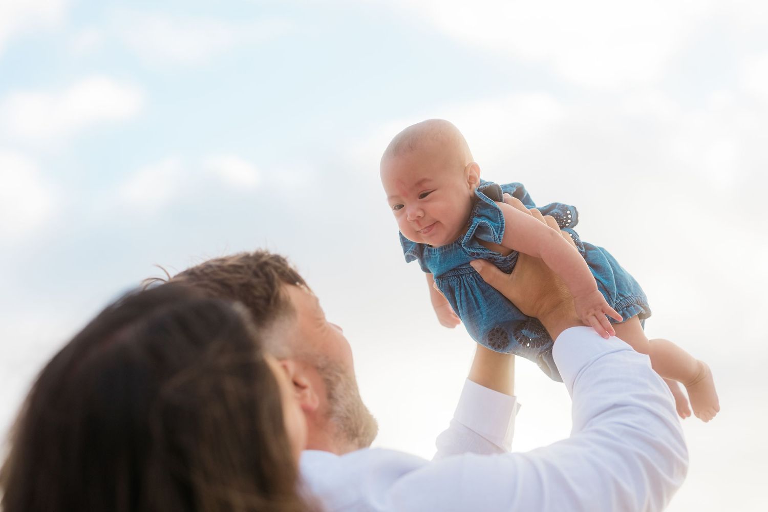 A joyful moment lifting a baby wearing blue against a bright sky background.