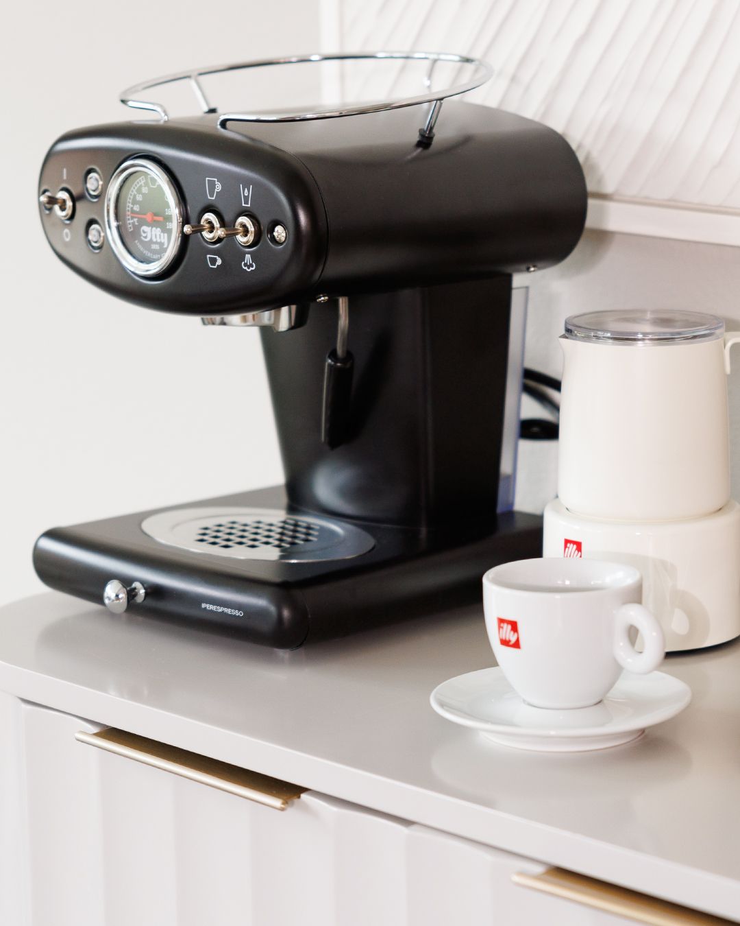 Modern black espresso machine setup with illy cups and saucers on white counter.
