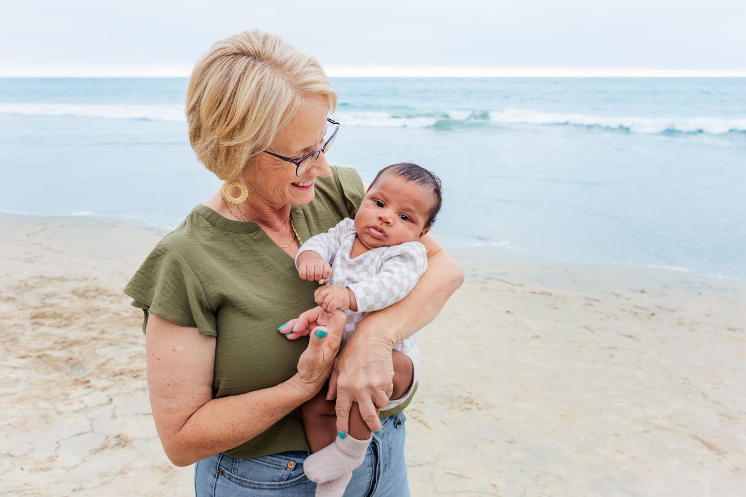 Adult holding small infant on sandy beach with ocean waves in background.