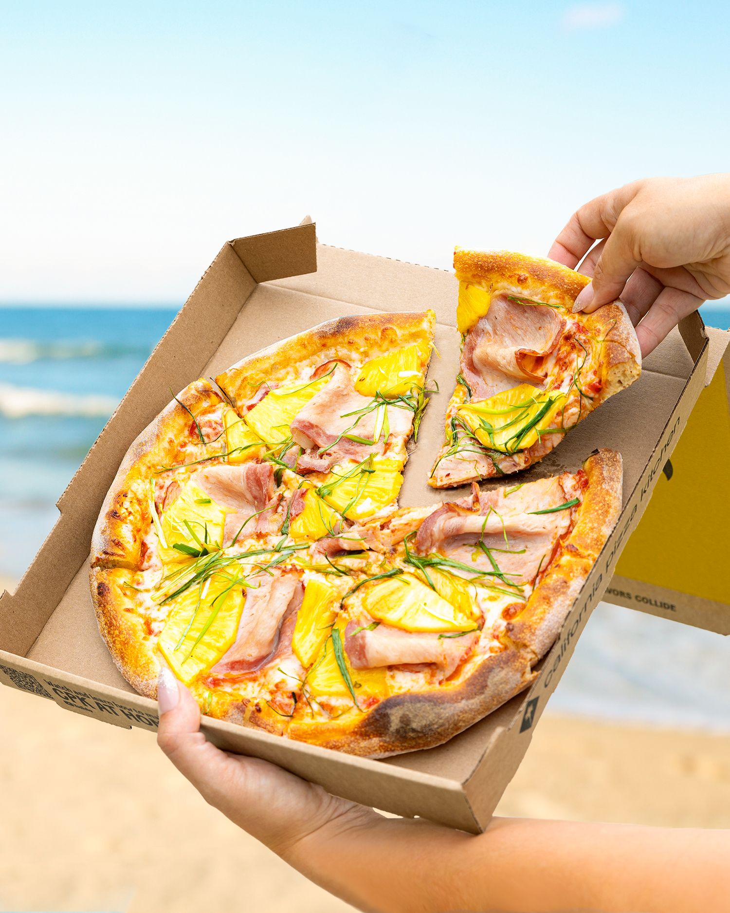 Pizza with fresh toppings being served in takeout boxes at the beach.