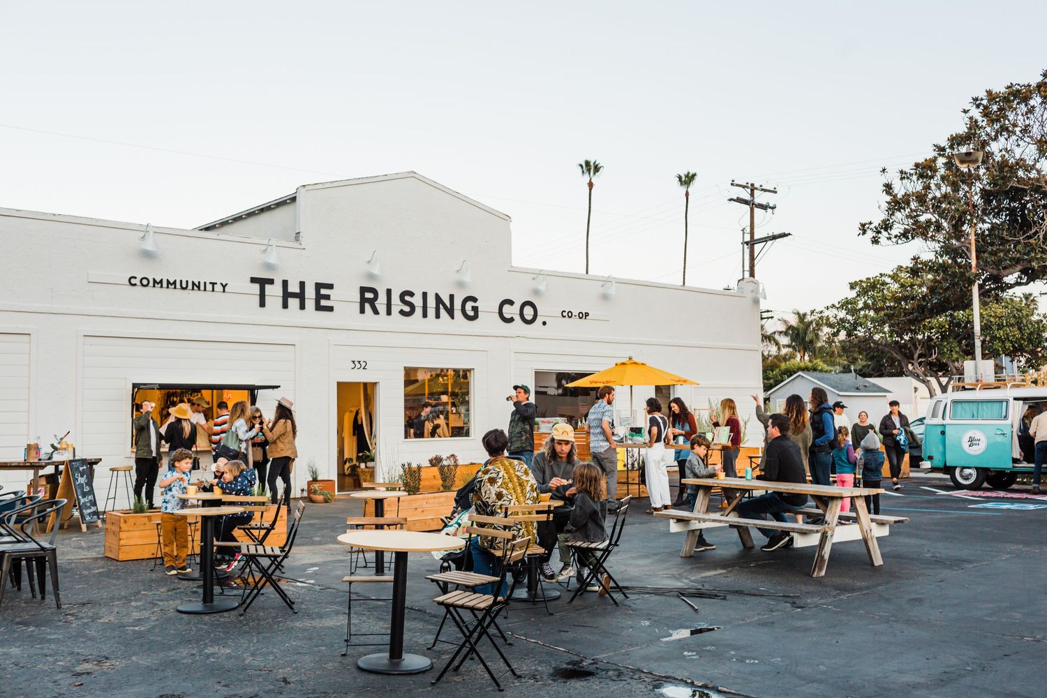 Customers gather at outdoor tables in front of The Rising Co. cafe building during a sunny day.
