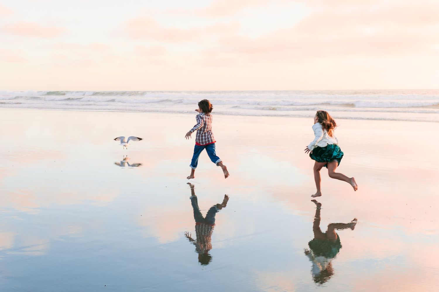 Two people run joyfully along a reflective beach at sunset casting shadows on the wet sand.