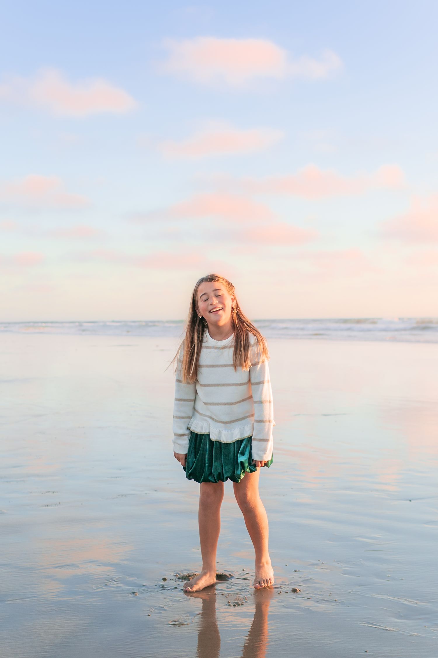 A child stands barefoot on a beach at sunset wearing a white sweater and green skirt against pastel colored clouds.