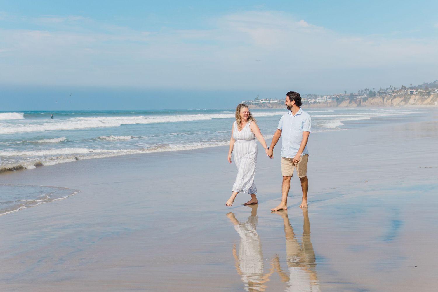 People walking along a scenic beach shoreline with waves and cloudy skies in the background.