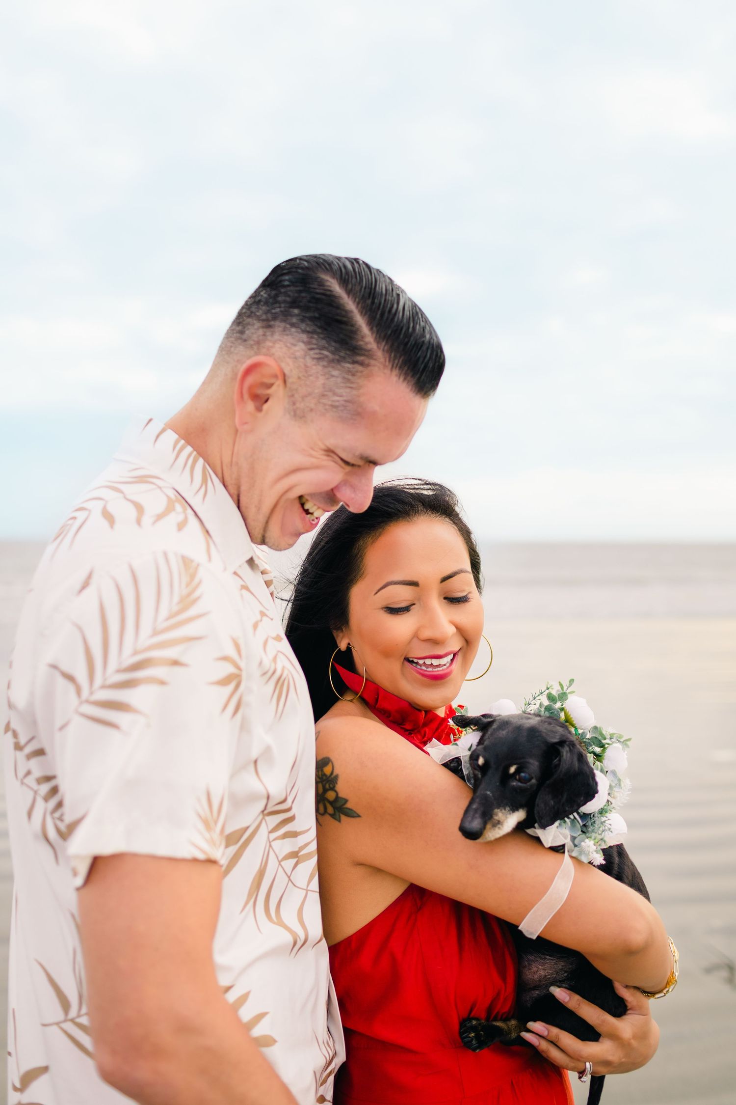 A couple in tropical attire shares a joyful moment while holding their small black dog on the beach.