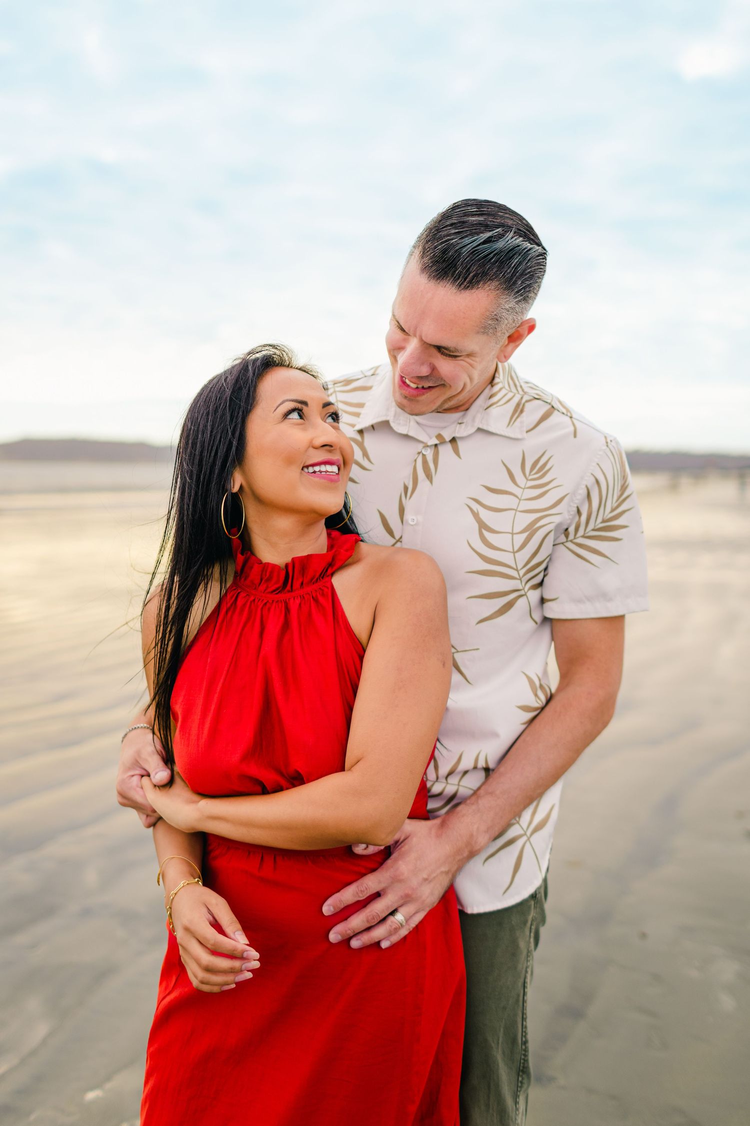 A couple shares a romantic moment on a sandy beach, with the woman wearing a vibrant red dress.