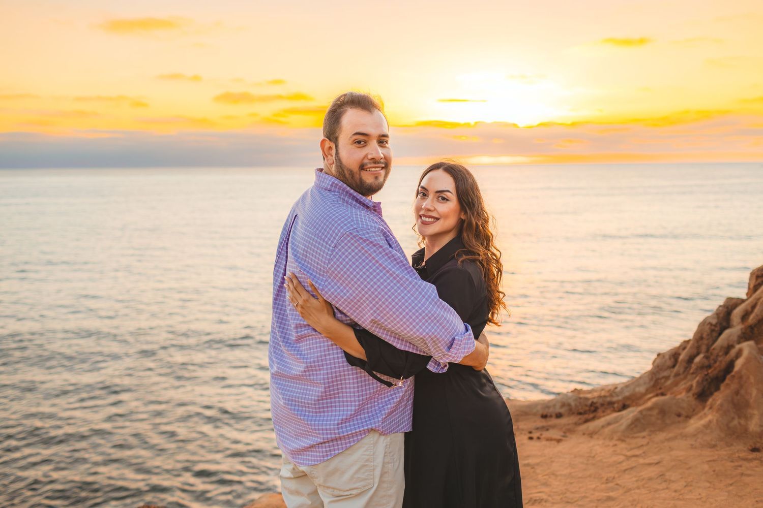 Couple embraces on coastal cliff during golden sunset over ocean waters.