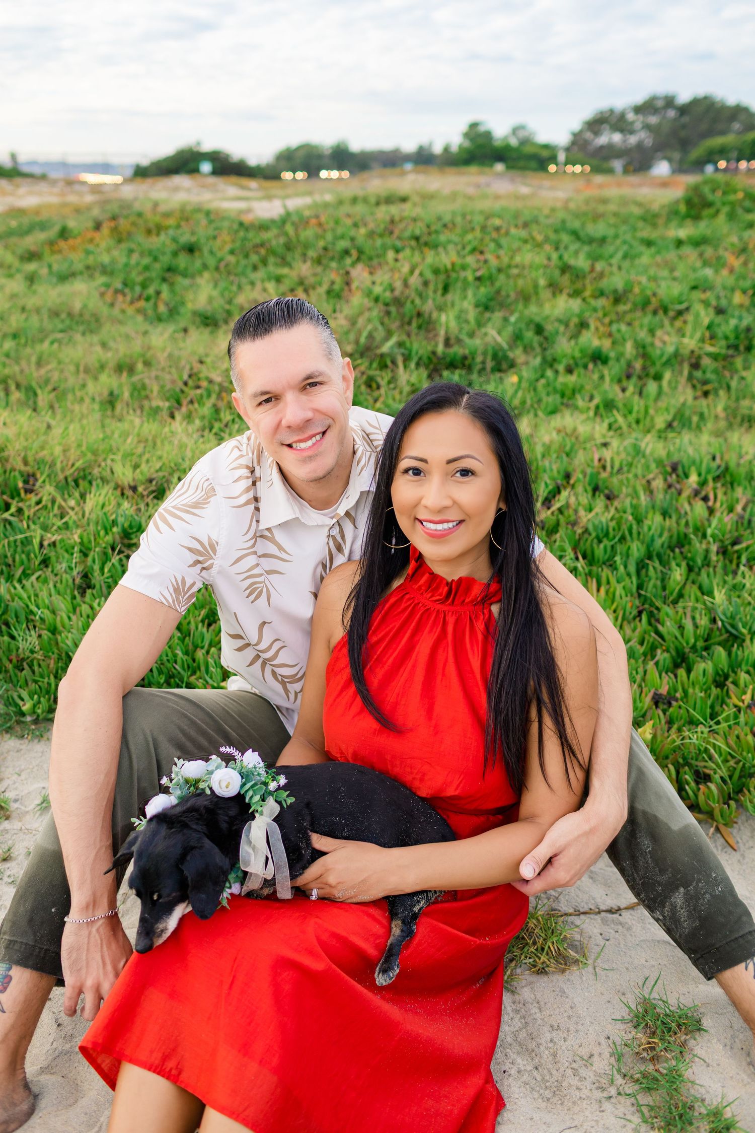 A couple sits together outdoors with their small black dog amidst green coastal vegetation.