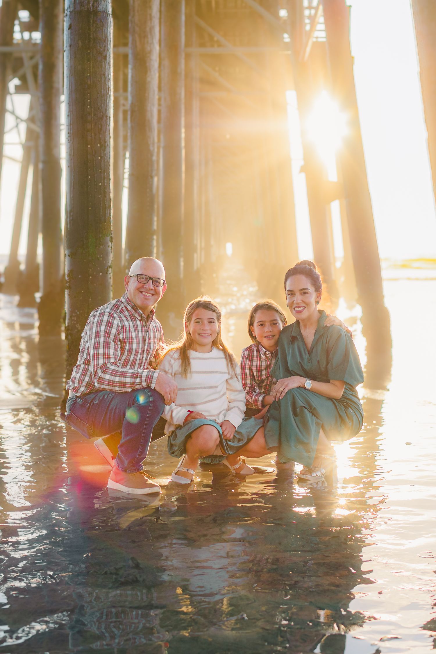 A family sits together under a pier at sunset with sunlight streaming through the wooden pillars.