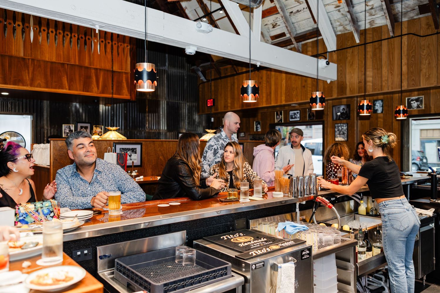Restaurant patrons enjoy drinks and food at a rustic wooden bar with pendant lighting and industrial decor.