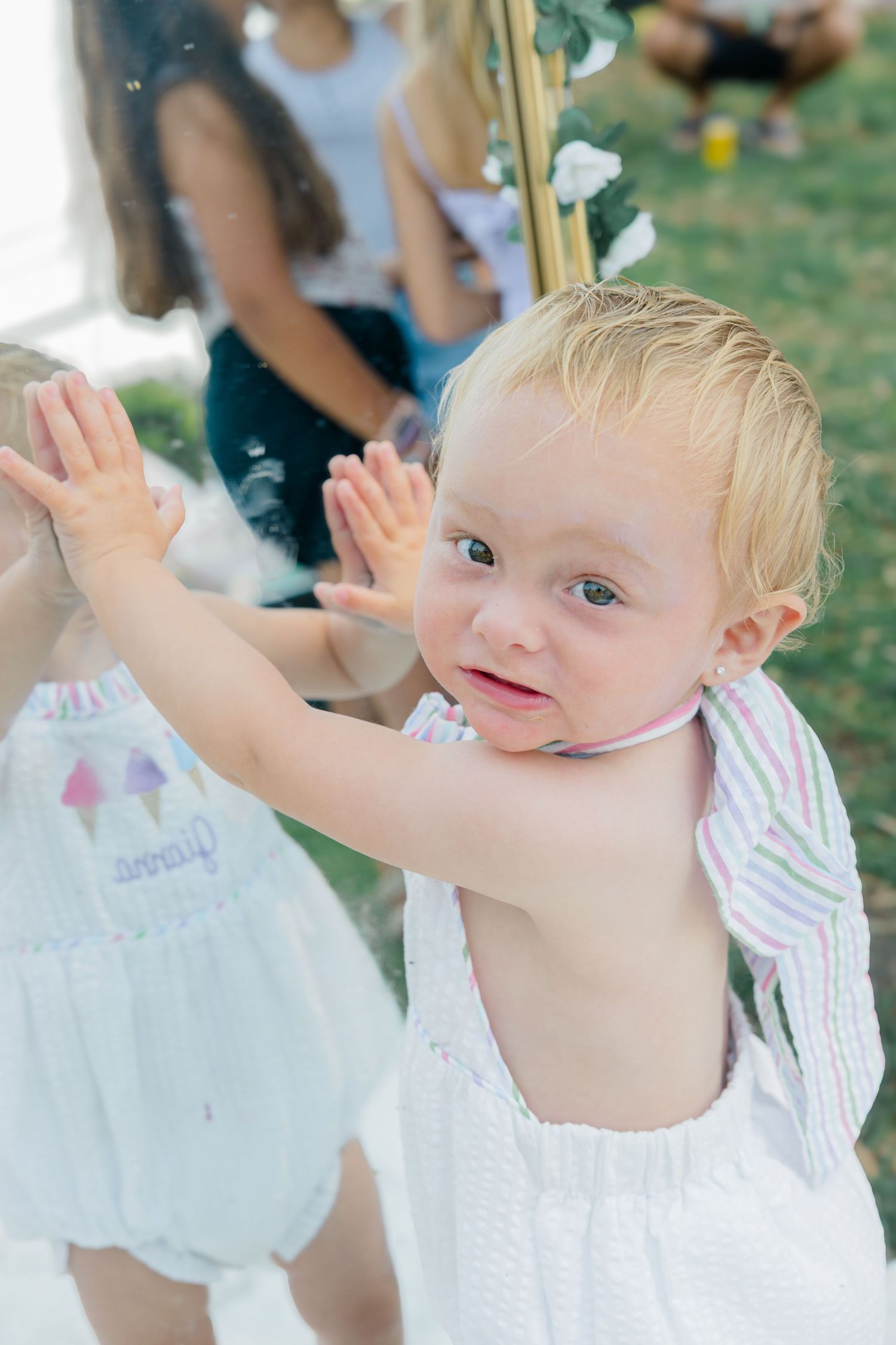 Baby in white dress playing and reaching up during outdoor summer photoshoot.
