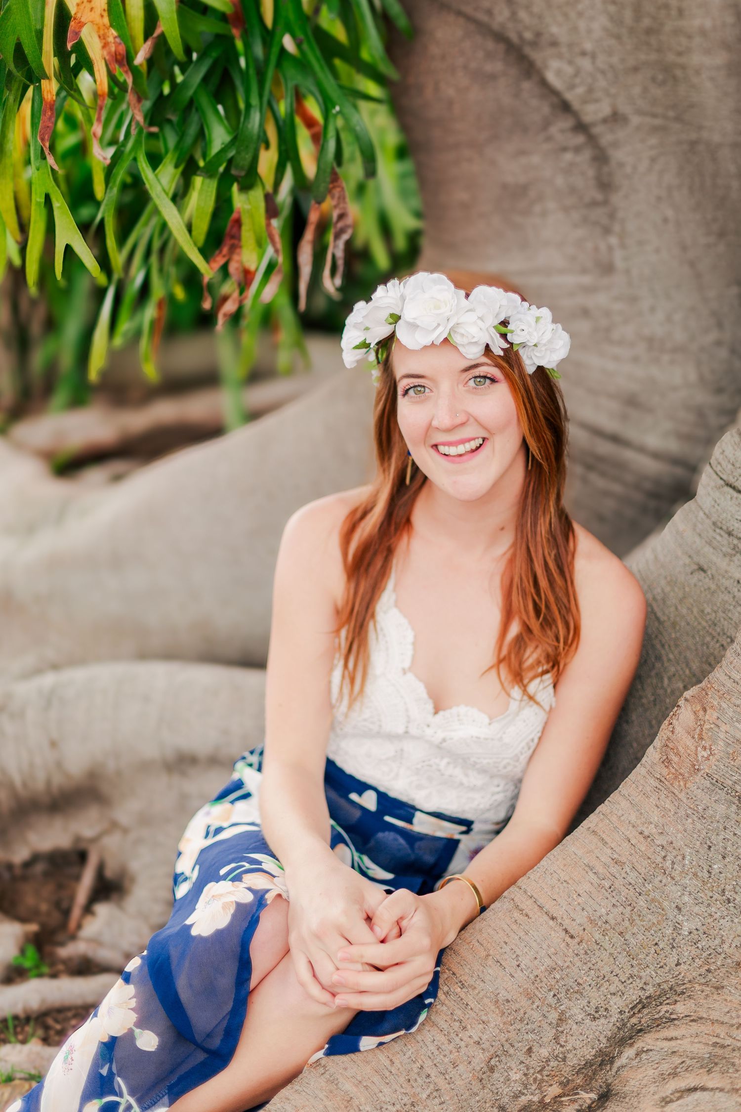 Someone wearing a white floral crown and lace top poses by a tree trunk in a blue floral skirt.