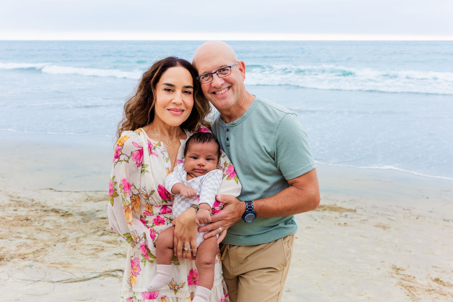 Three people share tender moment with infant on beach with waves in background.