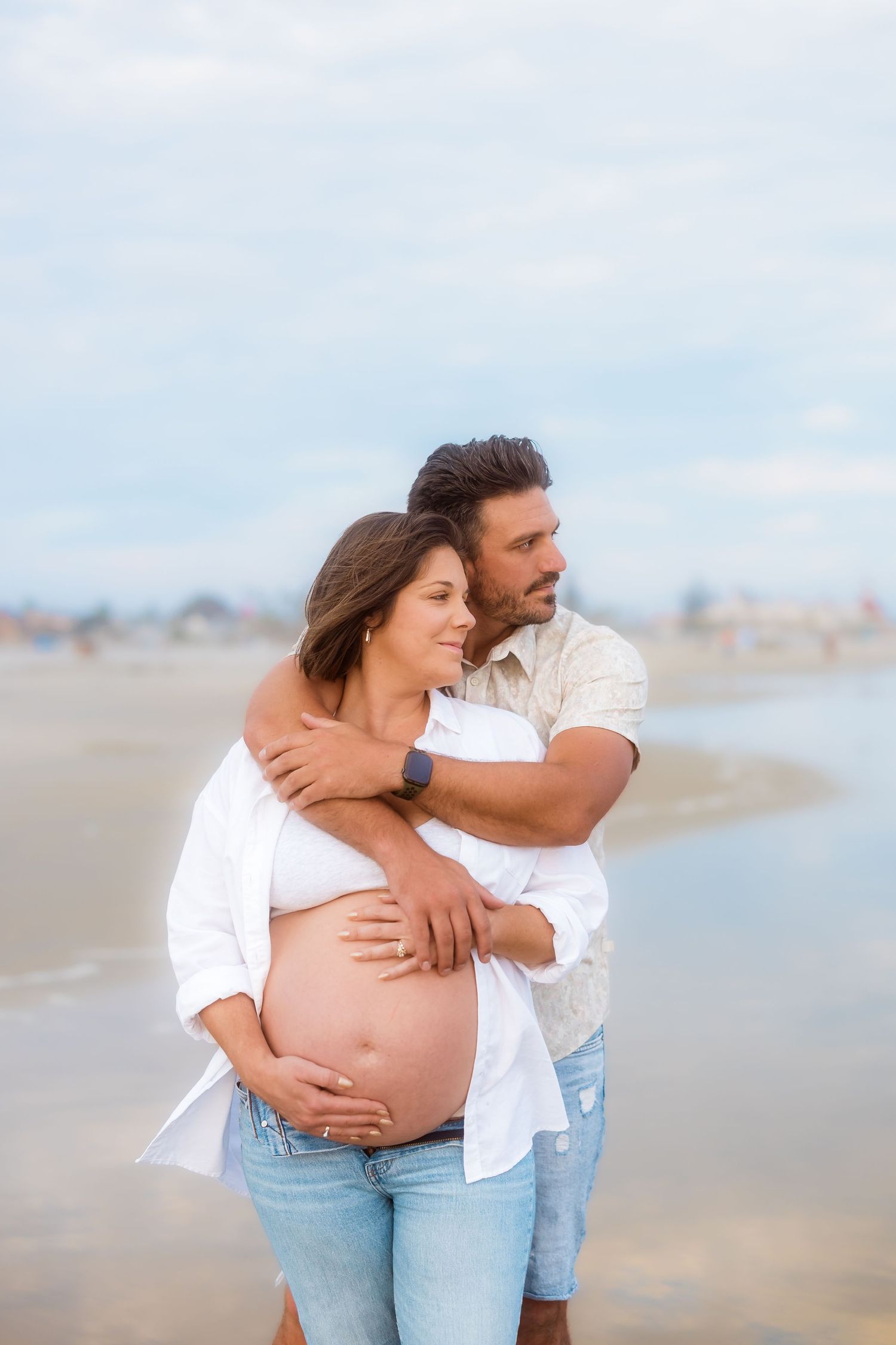 Intimate maternity photos of a couple embracing on a cloudy beach.