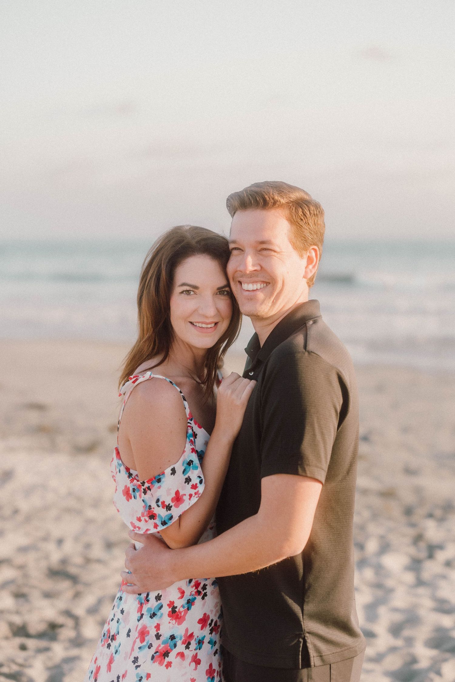 Romantic couple embraces on sandy beach at sunset in casual attire during engagement photoshoot.