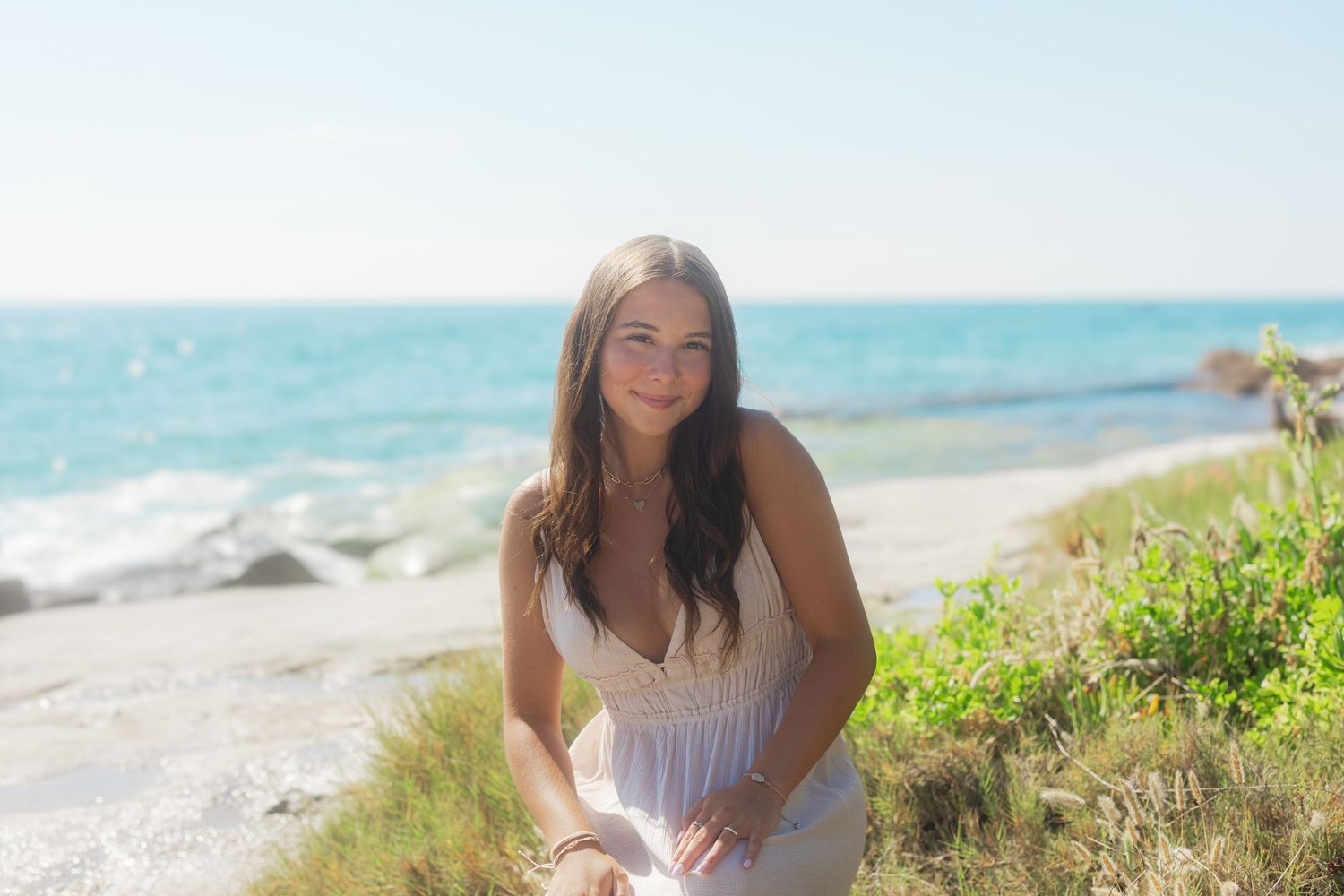 A woman in a white sundress poses on a sunny beach with turquoise ocean waves and coastal vegetation in the background.
