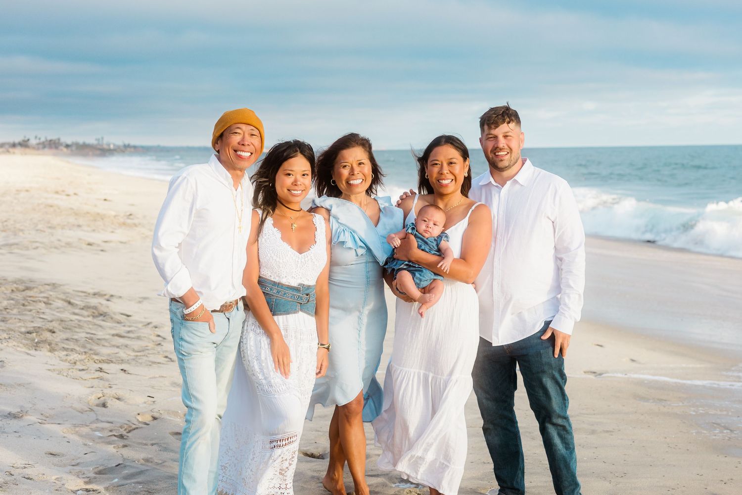 A family group poses together on the beach wearing coordinated white and blue outfits.