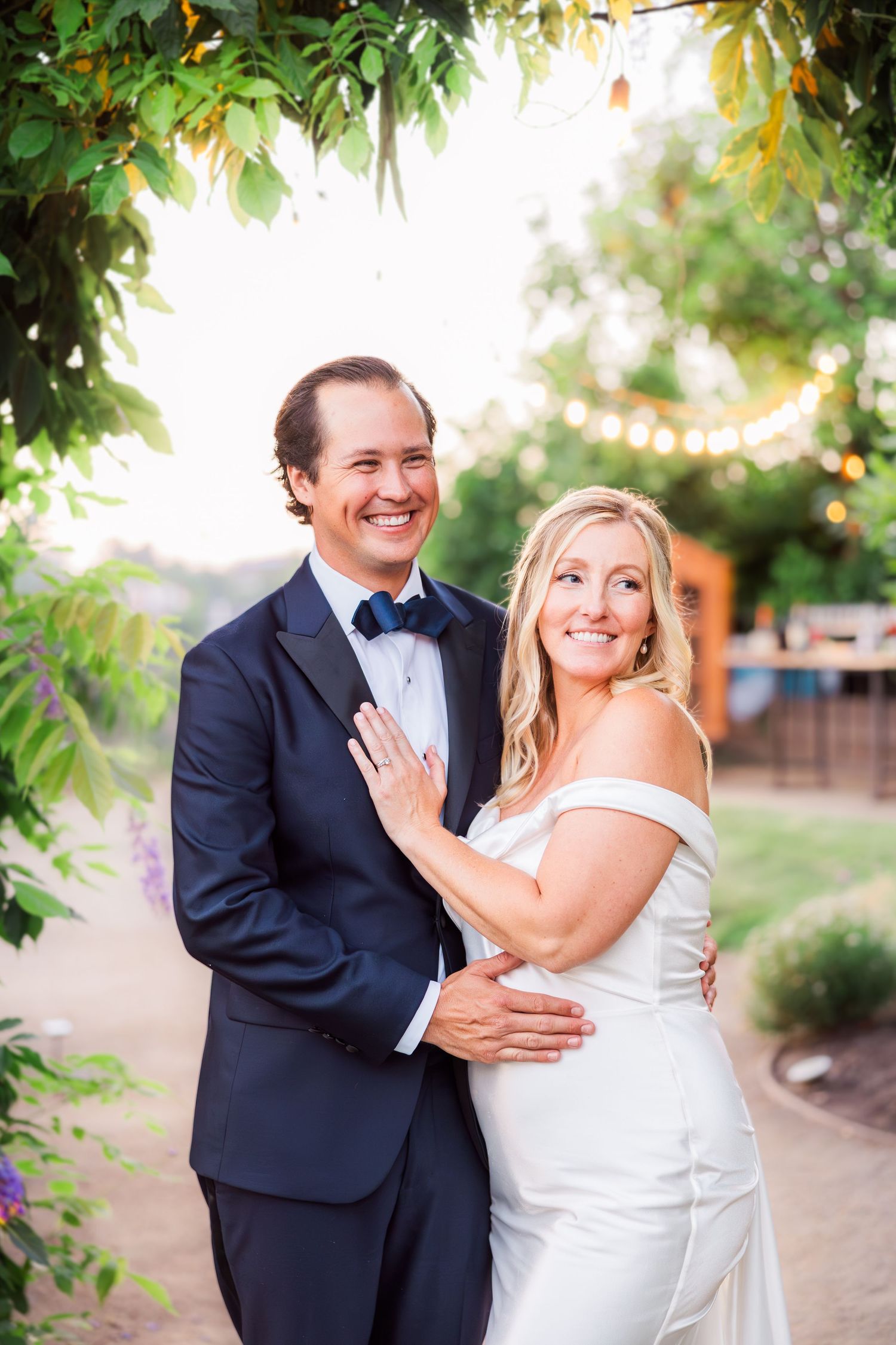 Joyful wedding couple laughing together in garden setting with string lights above.
