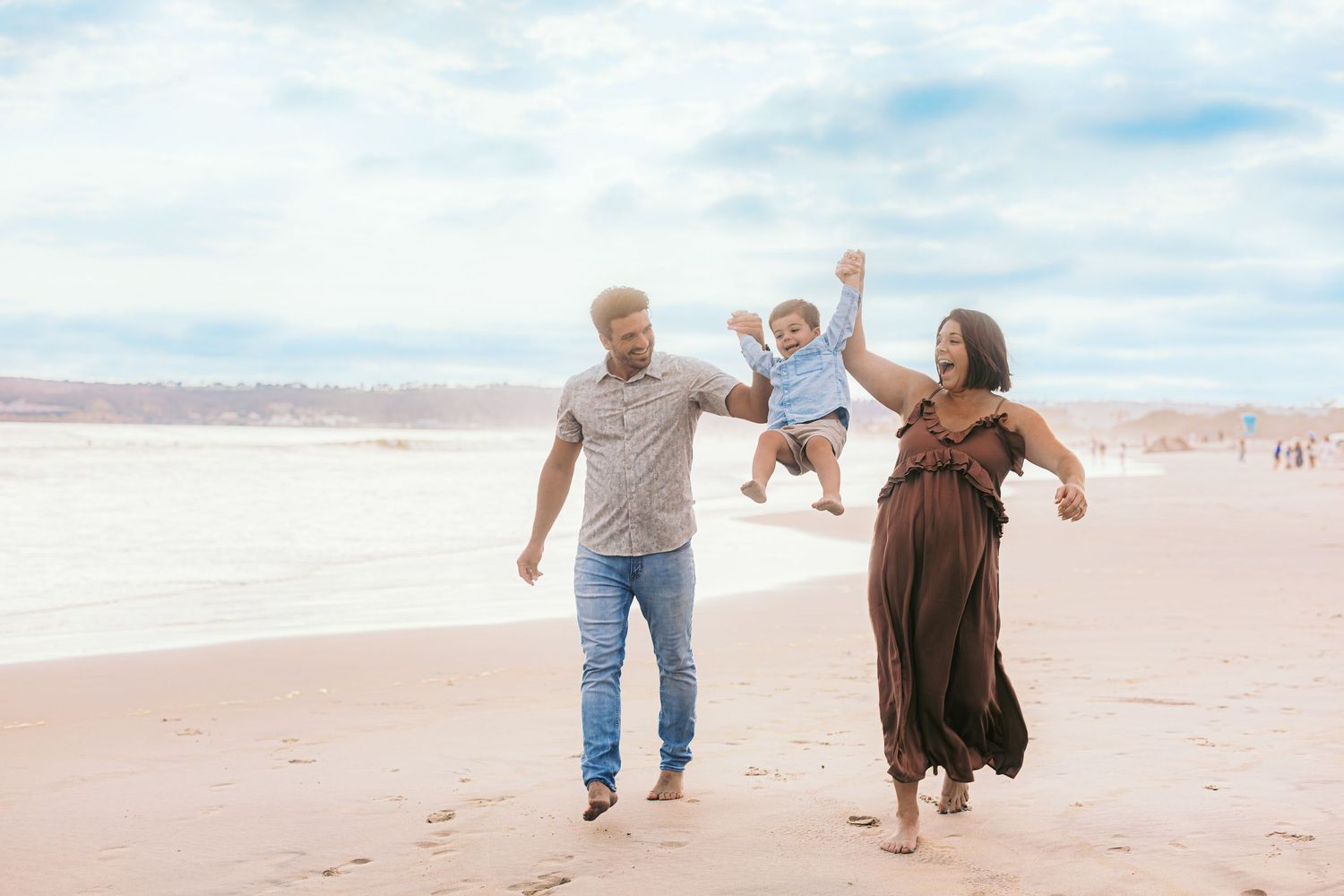 A family plays and swings their toddler while walking on the beach.