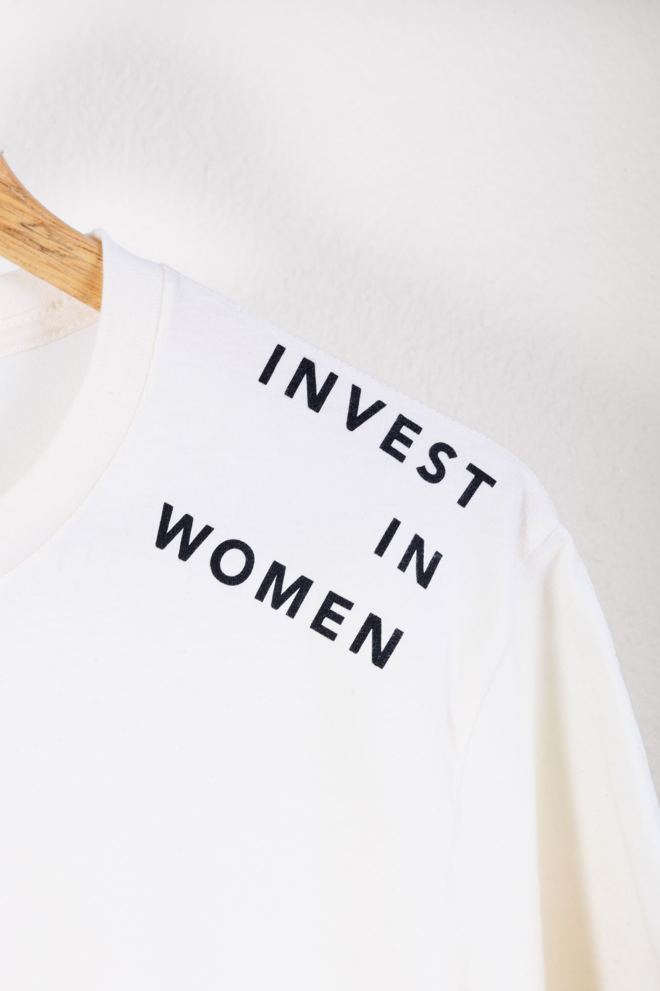 Close-up of white t-shirt with black text 'Invest in Women' on wooden hanger.