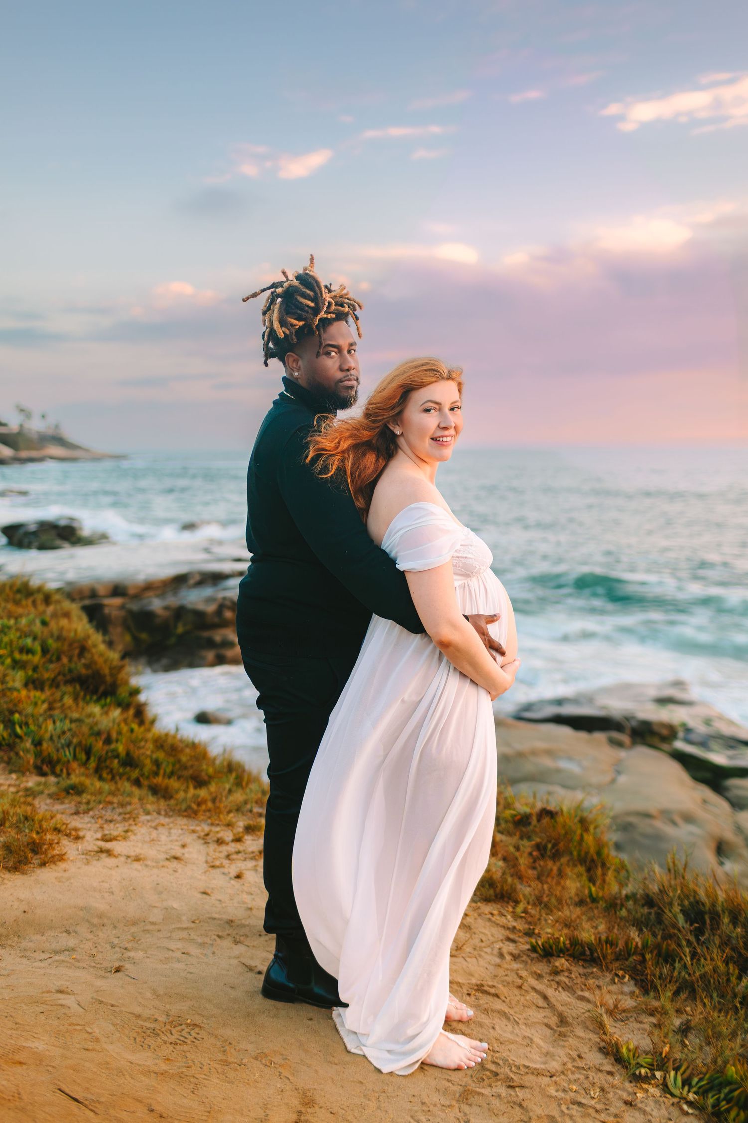 A couple stands together on a coastal cliff overlooking the ocean during a dreamy pink sunset.