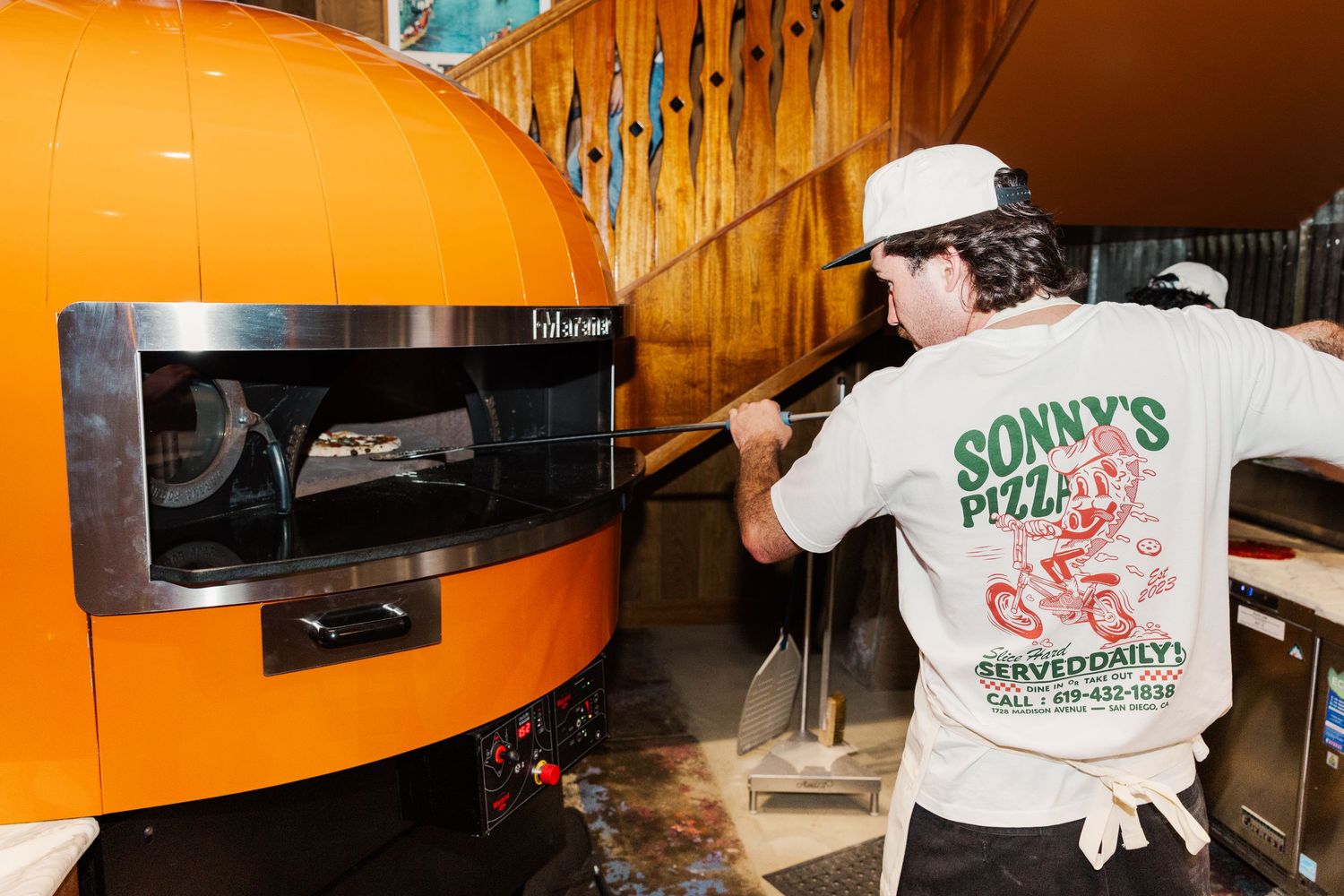 Restaurant worker operates an orange pizza oven while wearing a branded t-shirt.