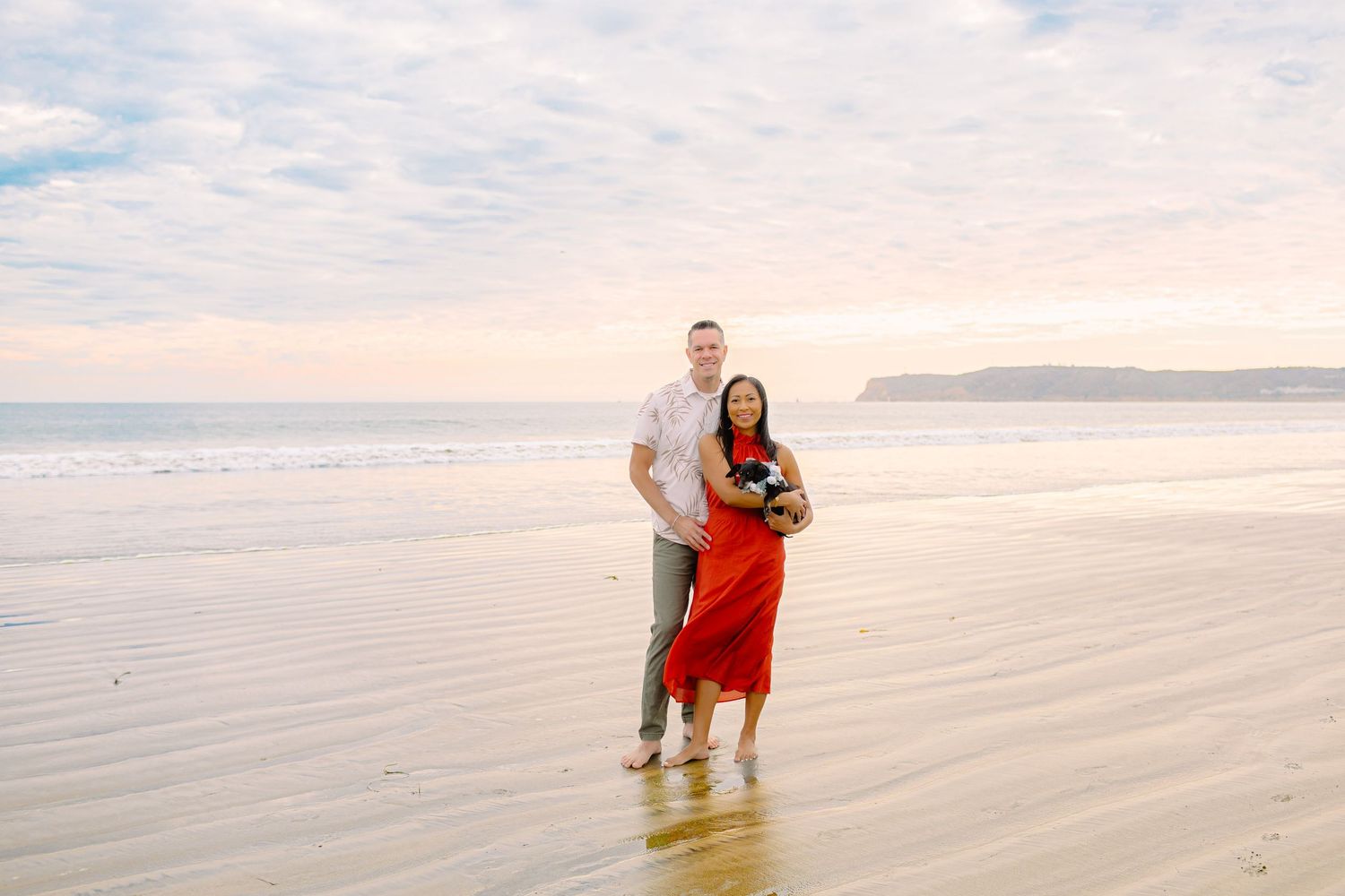 A couple stands together on a serene beach with white sand and cloudy skies in the background.