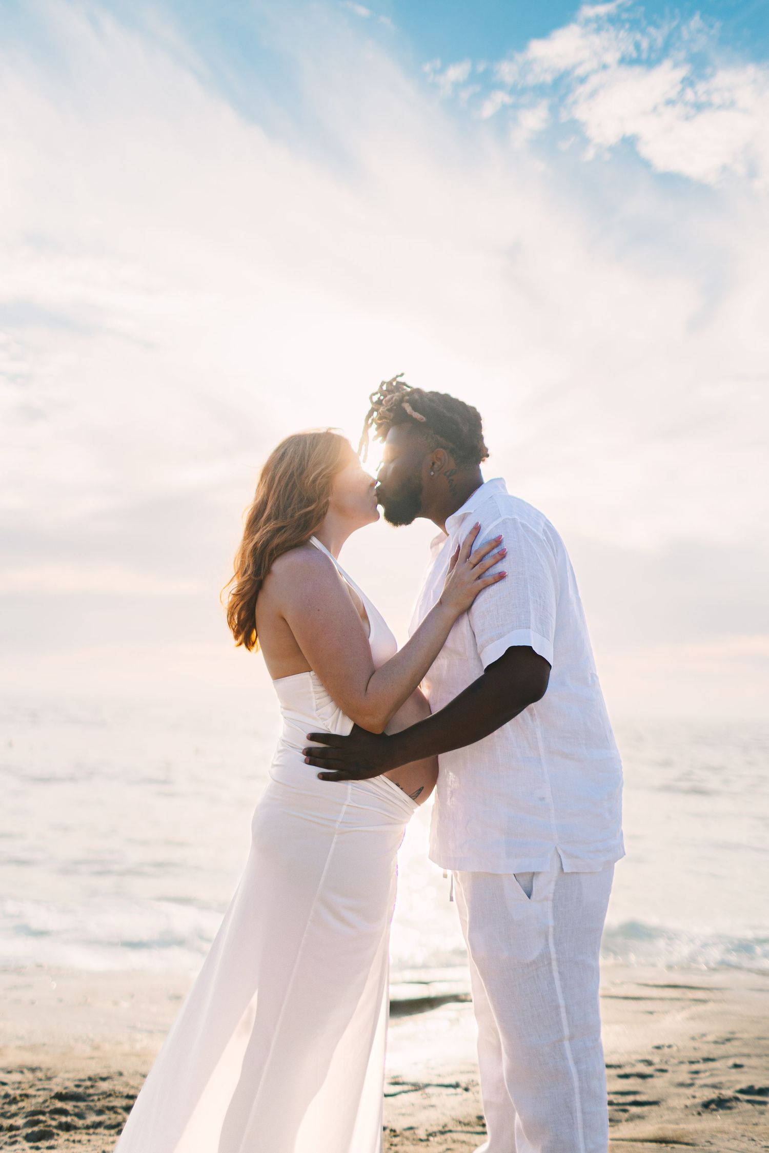 A couple in white attire embraces on a sunny beach with ocean waves and blue sky in the background.