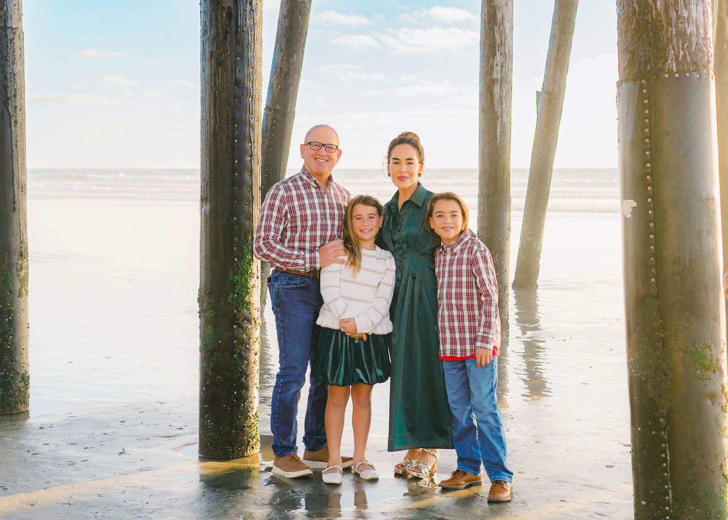 A family stands together on a beach with wooden pier pilings in the background during sunset.