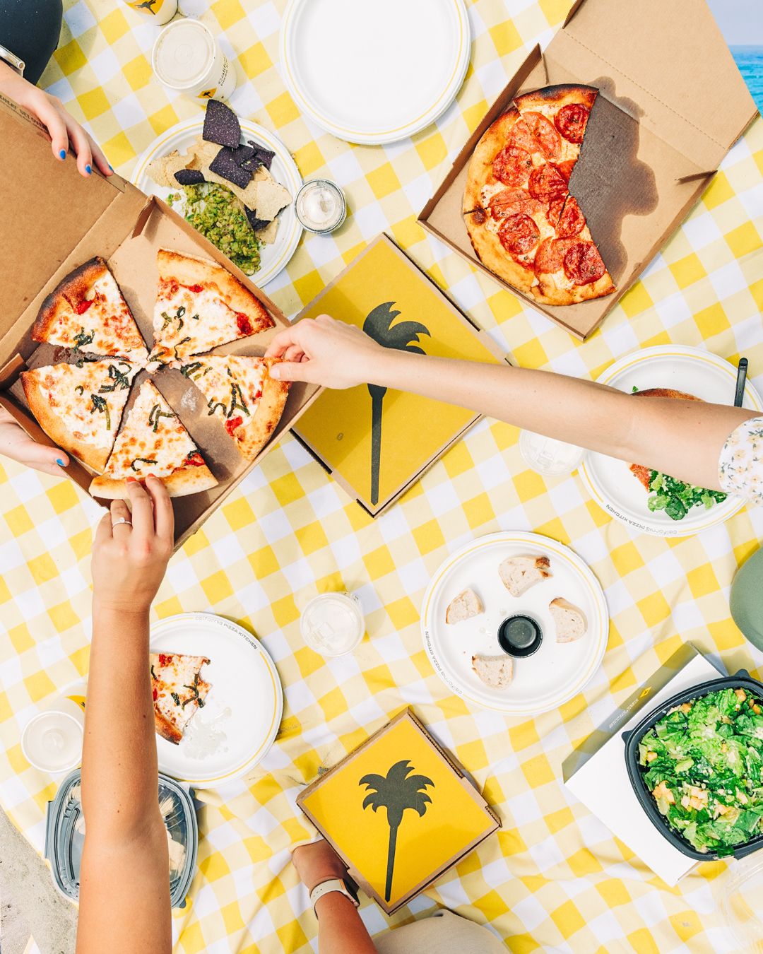 Hands reaching for pizza slices on yellow checkered tablecloth during beach picnic.
