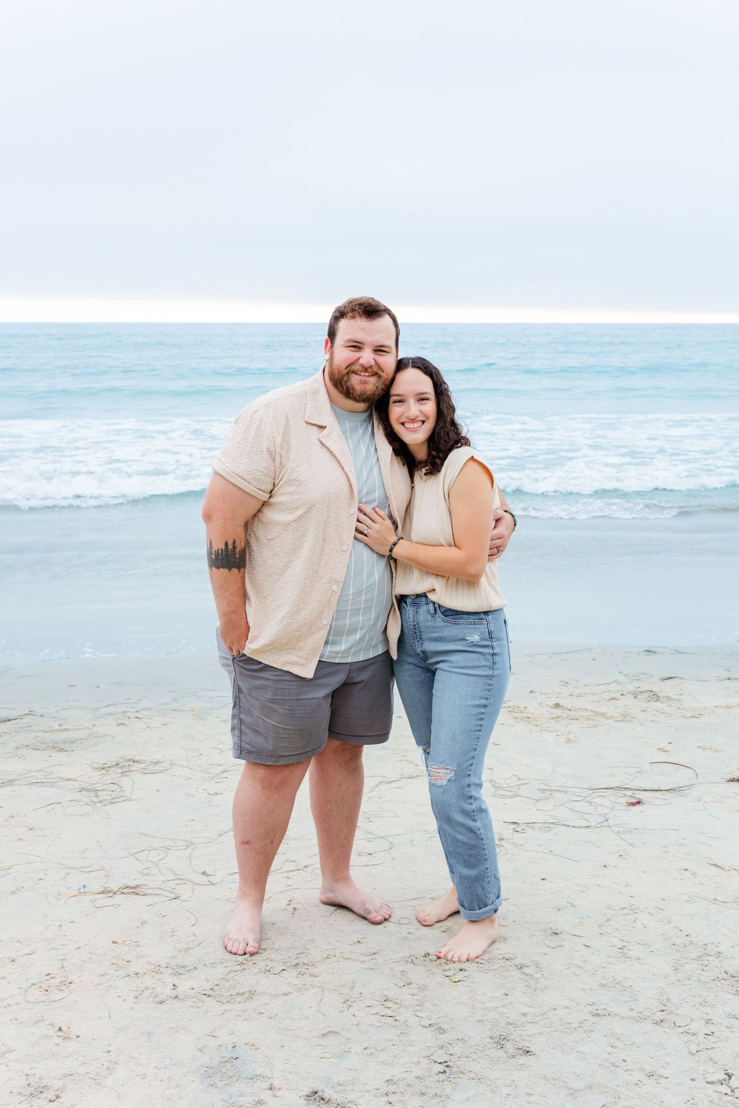 A couple stands barefoot together on the beach during an overcast day by the ocean.