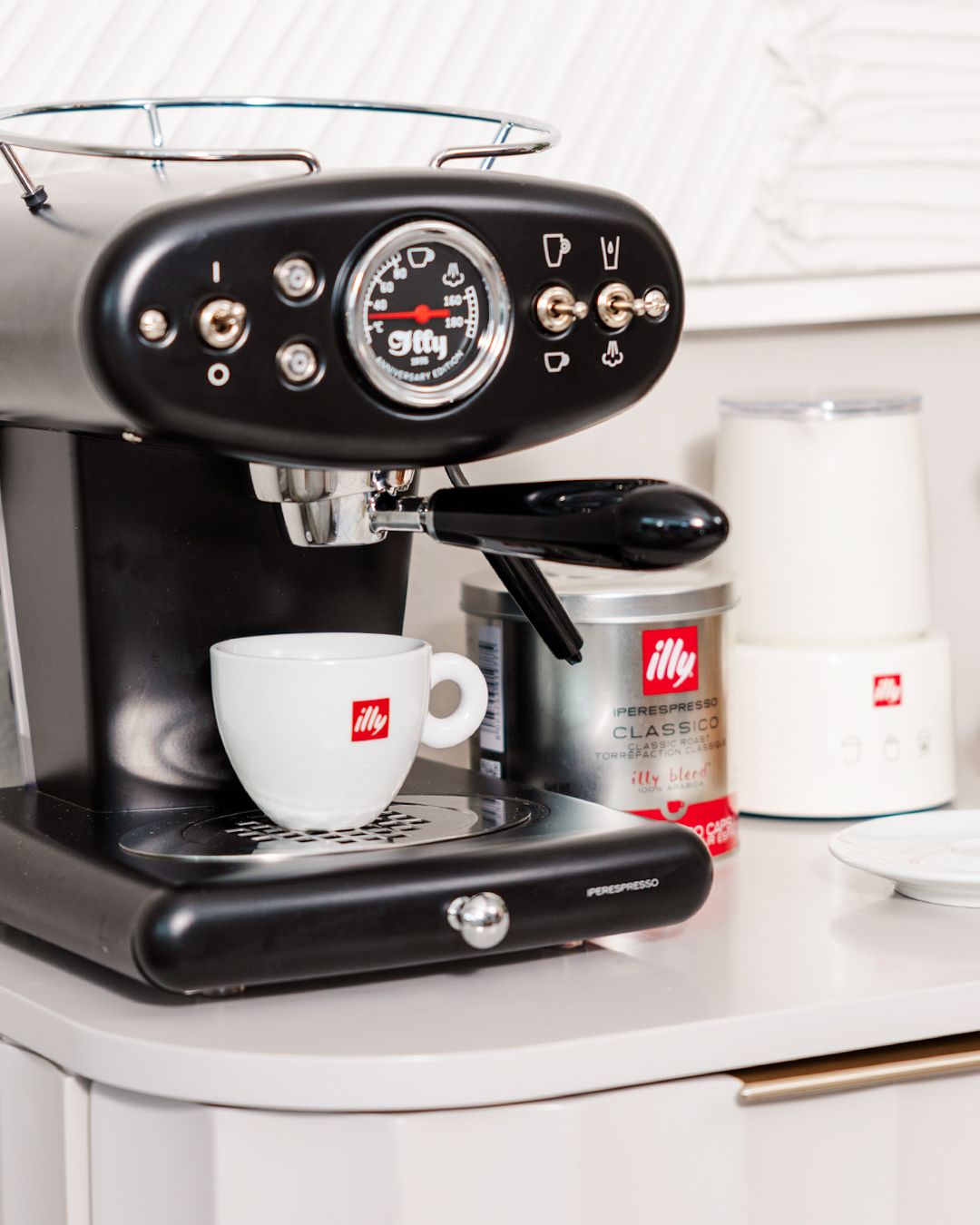 Black espresso machine with pressure gauge and illy coffee cups on white counter.