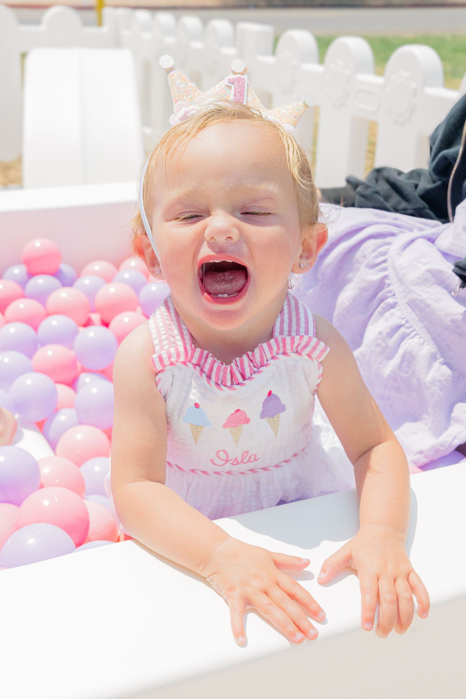 Laughing baby playing on white blanket surrounded by pink and purple balloons at outdoor party.