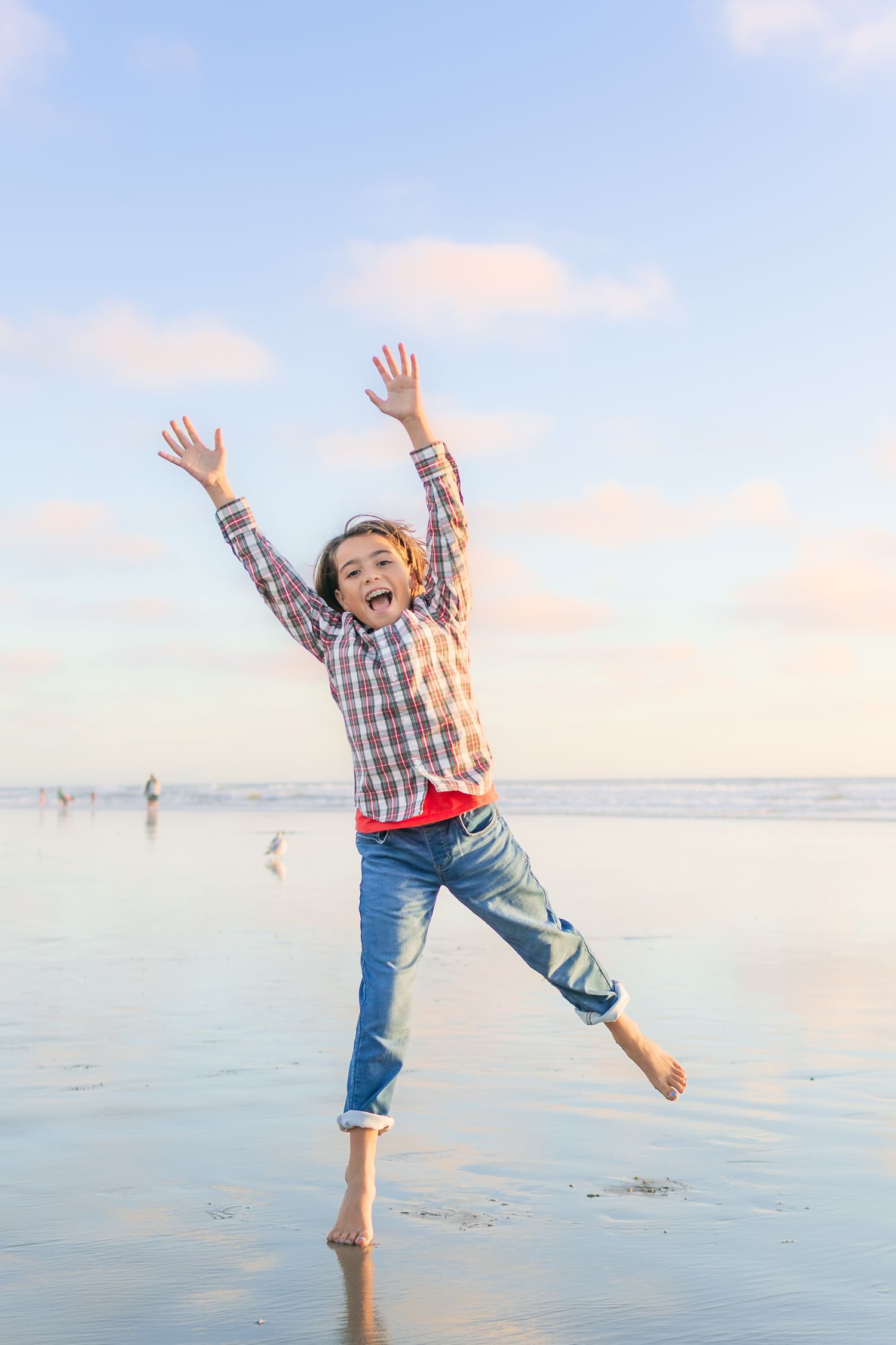 A person in a plaid shirt and jeans leaps joyfully on a beach at sunset with arms raised toward the sky.