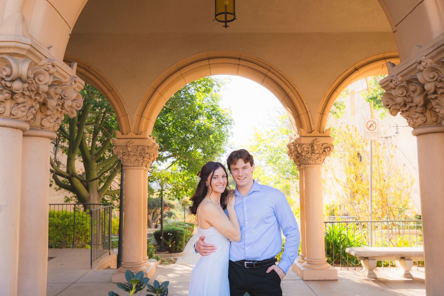 A couple stands together under ornate stone arches with sunlit gardens visible in the background.