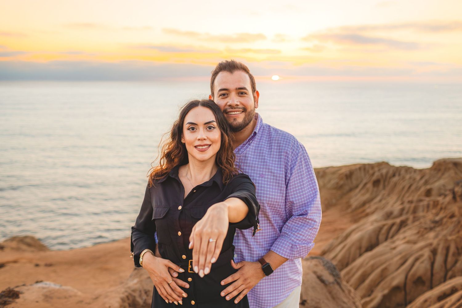 A couple embraces on a scenic coastal cliff during golden hour sunset by the ocean.